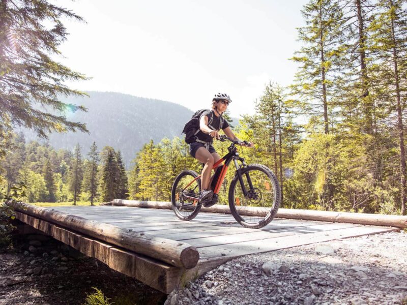 E-Bikerin fährt über eine Holzbrügge auf einem Wanderweg in den Bergen